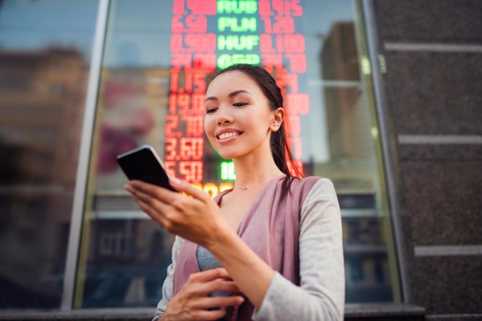 An investor, standing in front of an electronic board showing stock prices, looks at a phone.