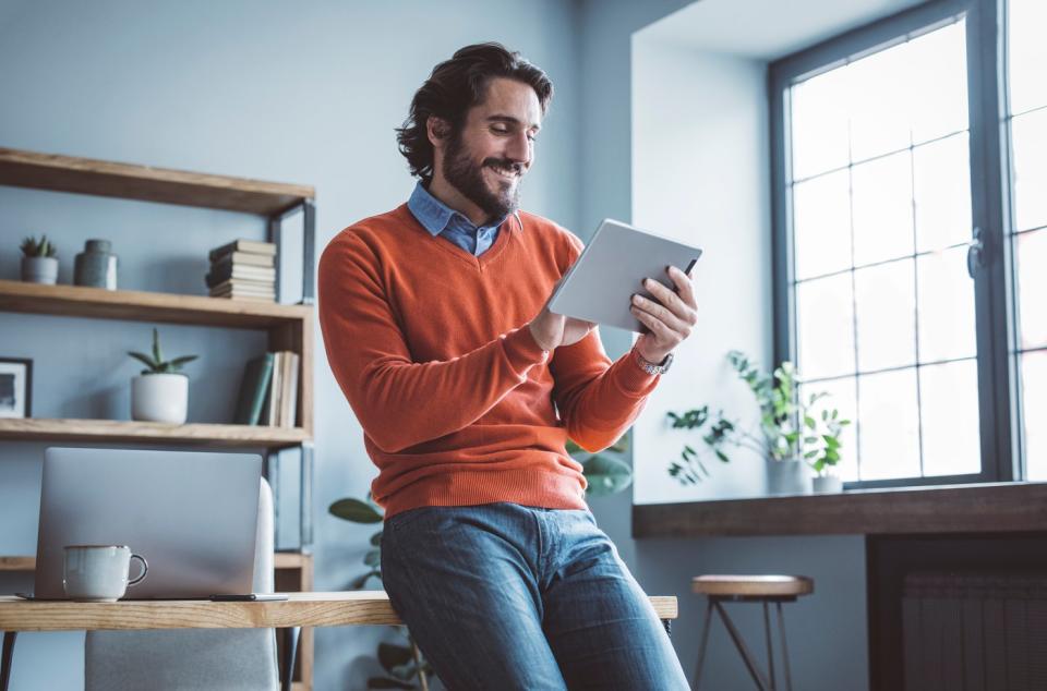 A smiling investor leans against a desk and looks at something on a tablet.