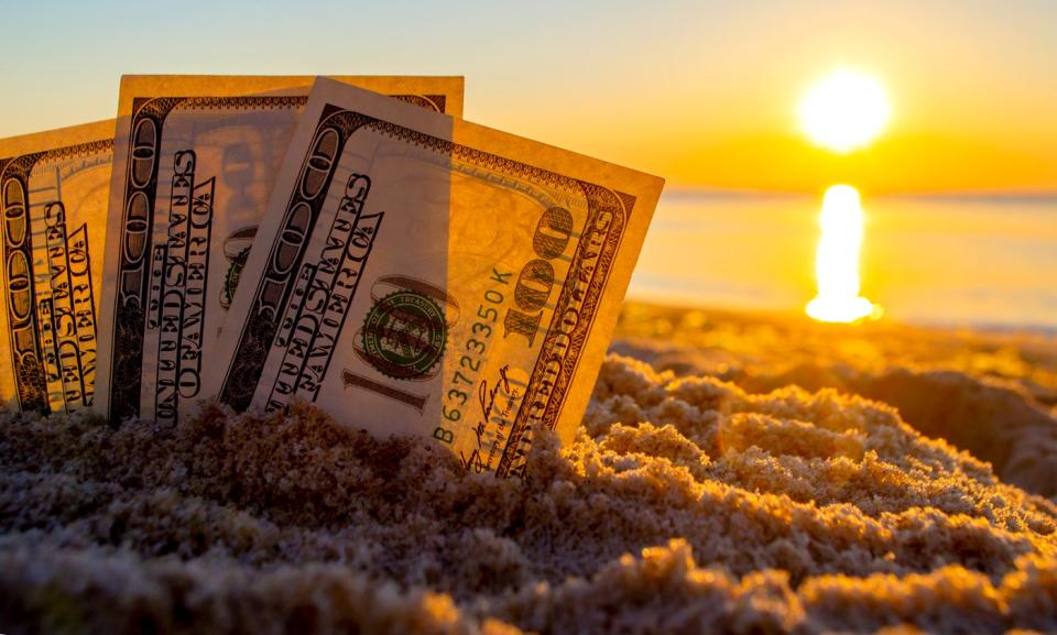 Three fanned one hundred dollar bills partially buried upright in the sand, with the sun in the background.